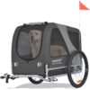 Ein Hund sitzt in einem grauen Doggyhut-Fahrradanhänger mit Netzfenstern und einer kleinen orangefarbenen Sicherheitsflagge.