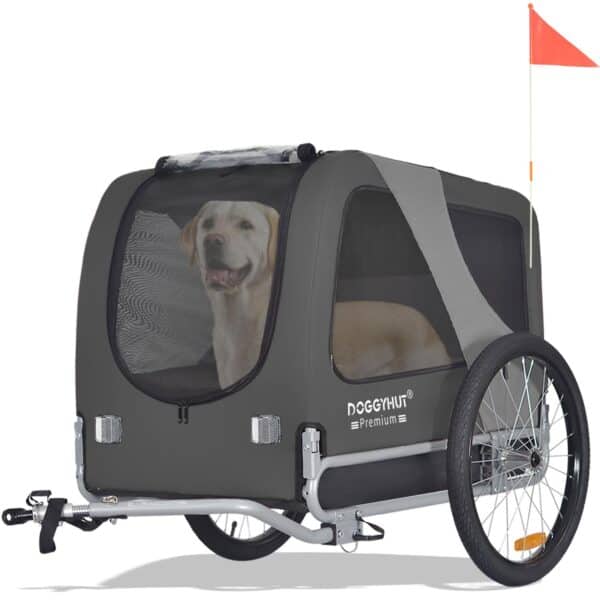 Ein Hund sitzt in einem grauen Doggyhut-Fahrradanhänger mit Netzfenstern und einer kleinen orangefarbenen Sicherheitsflagge.